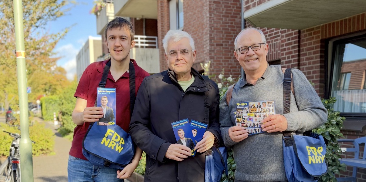 Drei Menschen mit FDP-Taschen und -Flyern vor Häusern